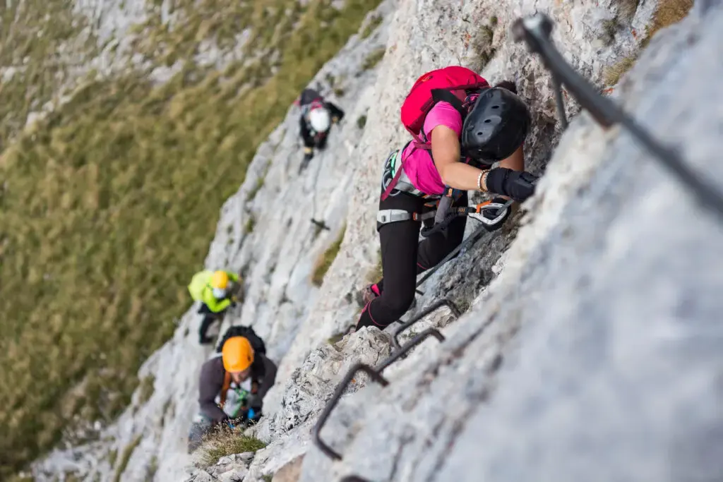 Woman and group of other people climbing on steep rock face on via ferrata. Climbers on via ferrata climbing route. Alpine ferrata. Summer adventure mountain activity_-4